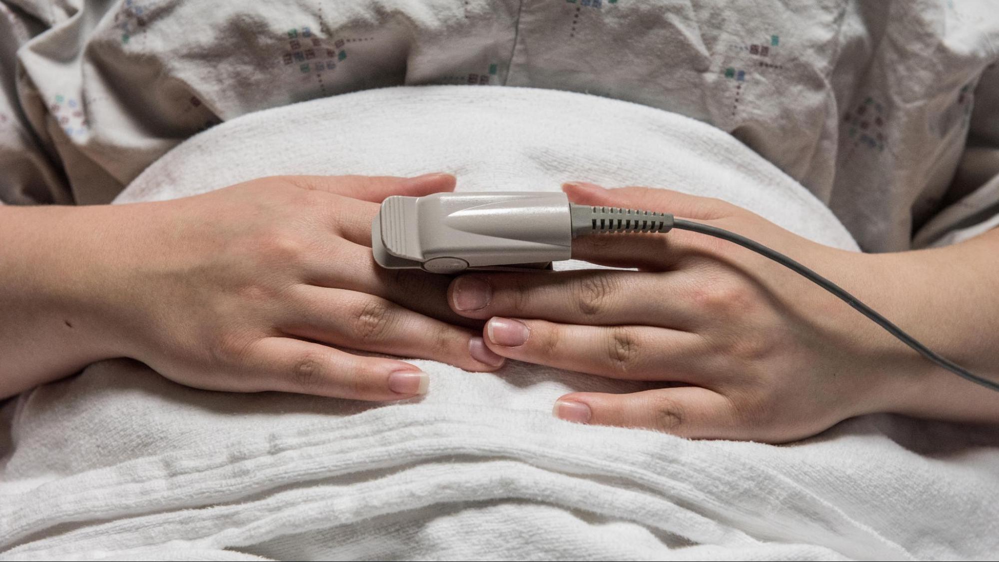 Patient in hospital bed with oxygen monitor attached to their pointer finger.